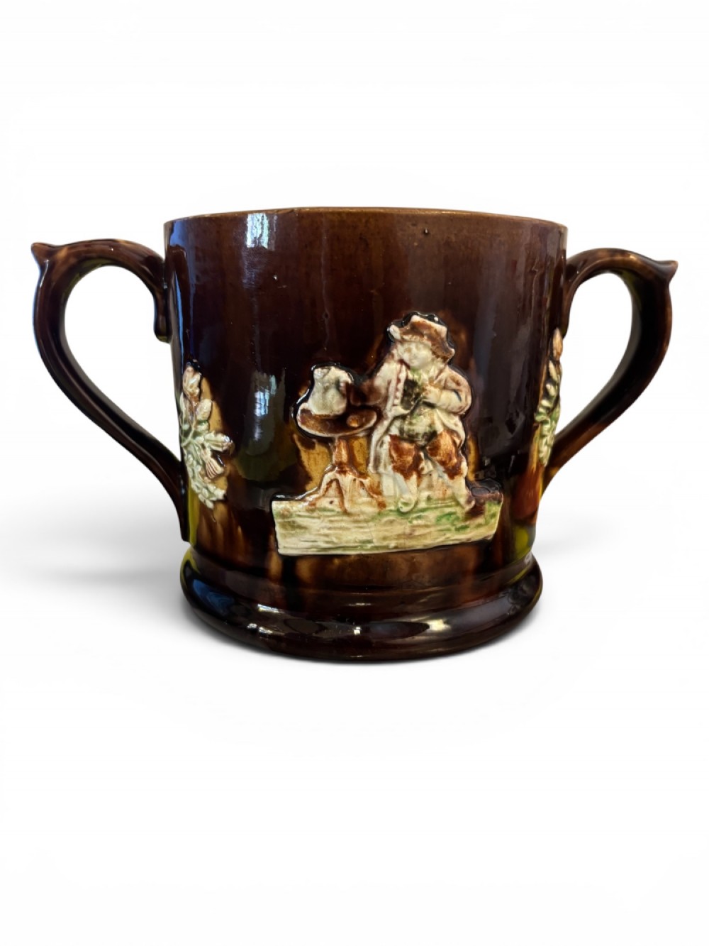 A dark brown glazed earthenware two-handled tankard featuring raised relief moulding of a seated figure drinking at a table.