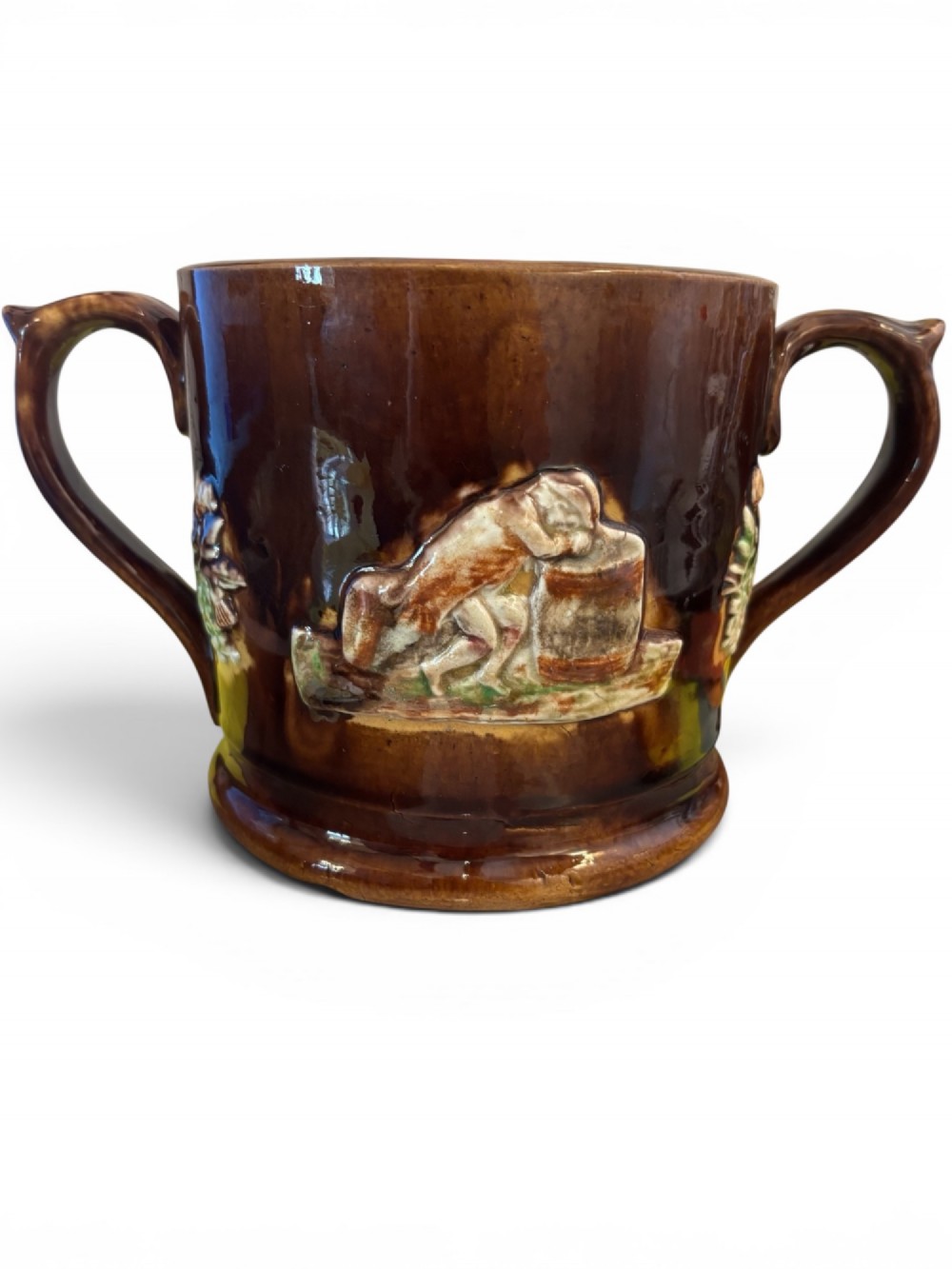 A dark brown glazed earthenware two-handled tankard featuring raised relief moulding of a seated figure drinking at a table. View II