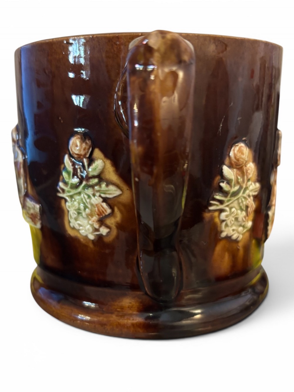 A dark brown glazed earthenware two-handled tankard featuring raised relief moulding of a seated figure drinking at a table. View III