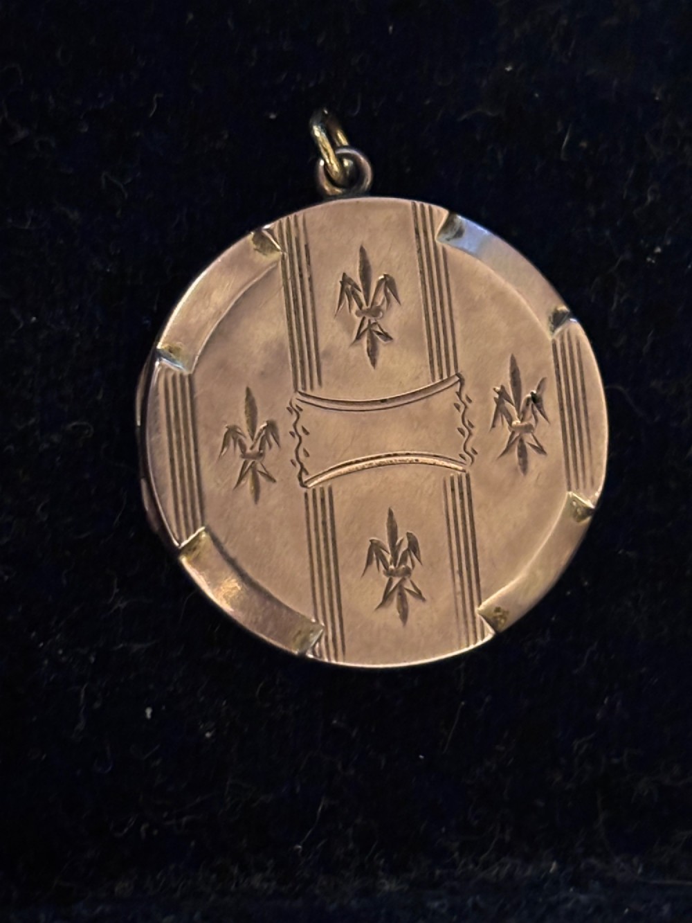 A circular antique gold locket featuring hand-engraved fleur-de-lis emblems and vertical geometric stripes surrounding an unengraved central cartouche.