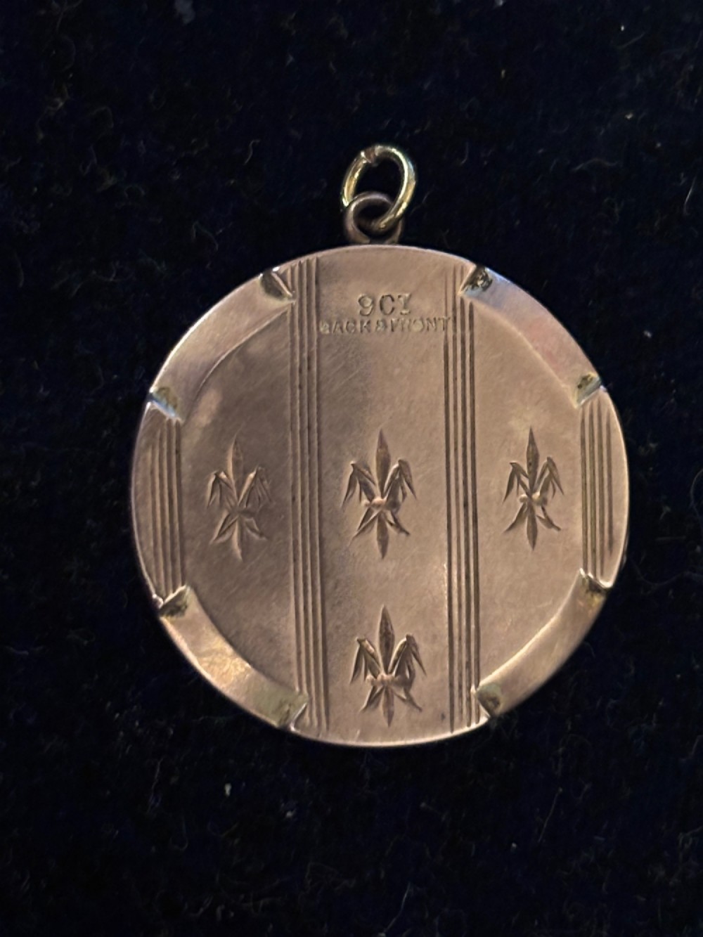 A circular antique gold locket featuring hand-engraved fleur-de-lis emblems and vertical geometric stripes surrounding an unengraved central cartouche. View IV