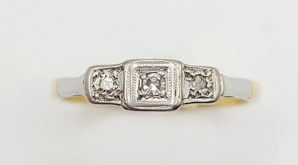 An Art Deco platinum-set and 18ct gold trilogy diamond ring featuring three central stones within a geometric bezel and millgrained border.
