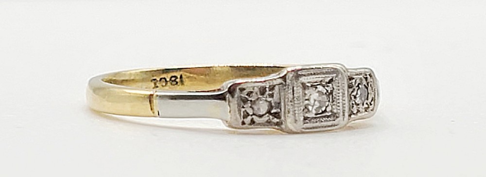 An Art Deco platinum-set and 18ct gold trilogy diamond ring featuring three central stones within a geometric bezel and millgrained border. View II