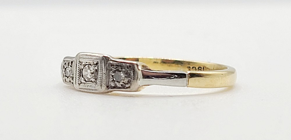 An Art Deco platinum-set and 18ct gold trilogy diamond ring featuring three central stones within a geometric bezel and millgrained border. View IV