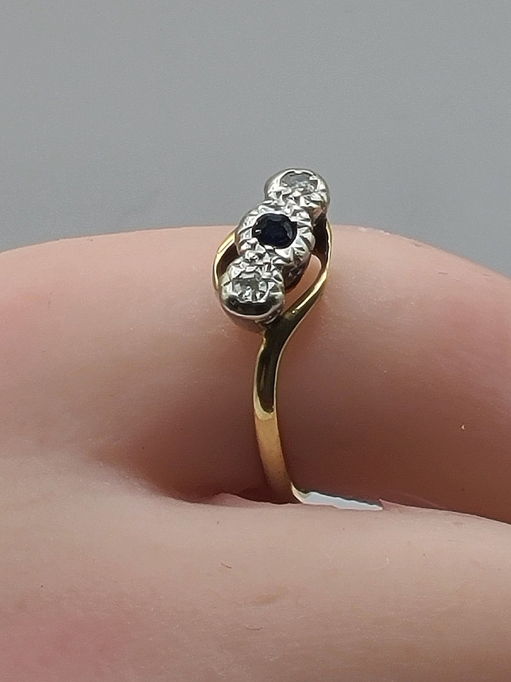An Art Deco trilogy twist ring featuring a central round blue sapphire flanked by two diamonds set in platinum atop an 18ct yellow gold band. View III