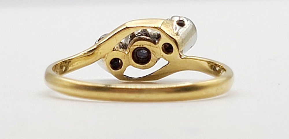 An Art Deco trilogy twist ring featuring a central round blue sapphire flanked by two diamonds set in platinum atop an 18ct yellow gold band. View V