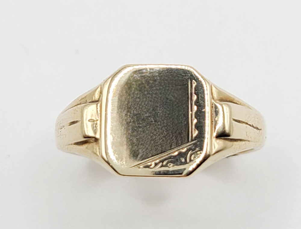 A close-up view of an antique 9ct gold art deco signet ring featuring geometric engraving on the shoulders and a rectangular face.