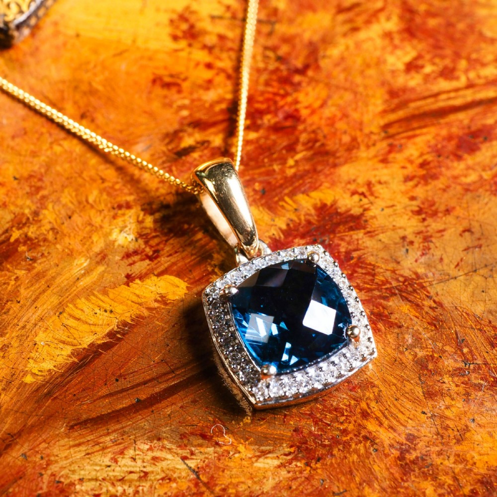 A cushion-cut deep blue topaz gemstone set in a square diamond halo pendant on a gold chain. View II