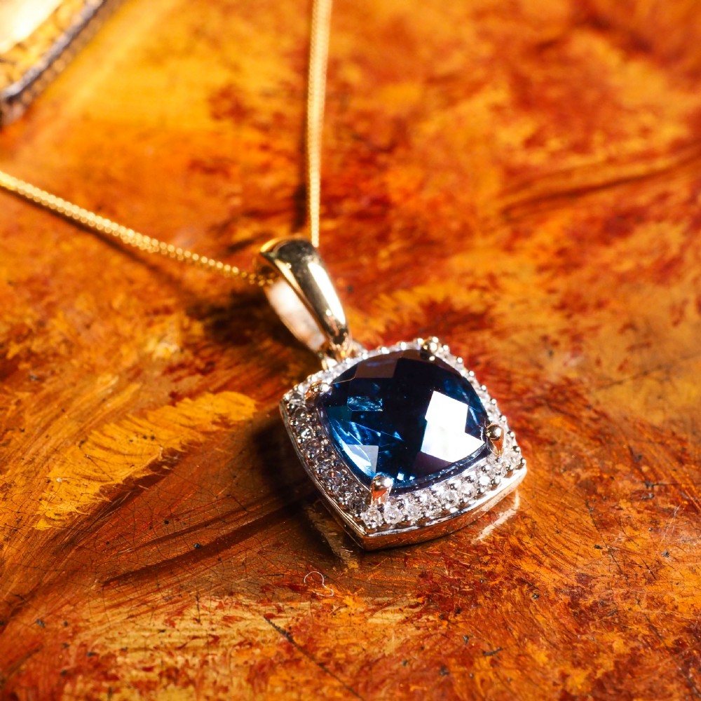 A cushion-cut deep blue topaz gemstone set in a square diamond halo pendant on a gold chain. View III