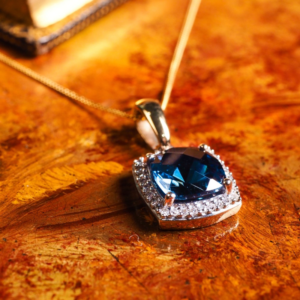 A cushion-cut deep blue topaz gemstone set in a square diamond halo pendant on a gold chain. View VI