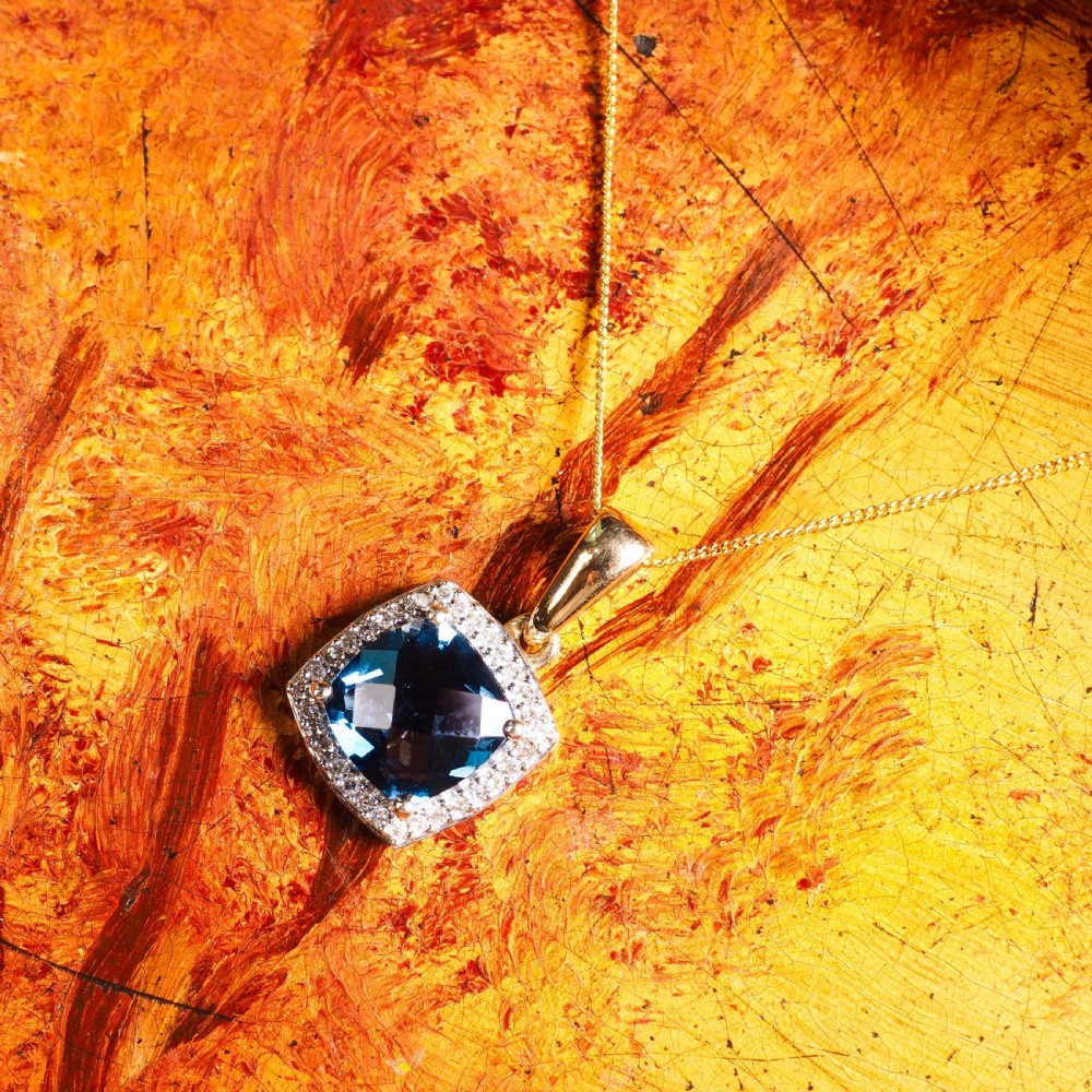 A cushion-cut deep blue topaz gemstone set in a square diamond halo pendant on a gold chain. View VIII