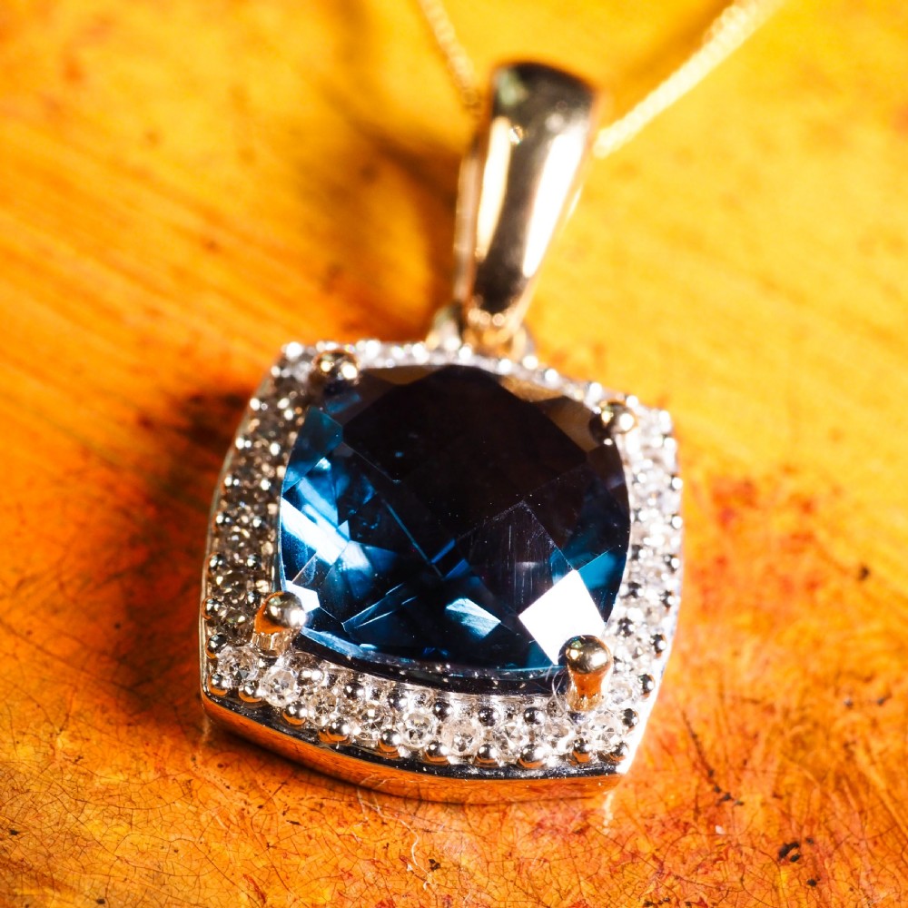 A cushion-cut deep blue topaz gemstone set in a square diamond halo pendant on a gold chain. View IX