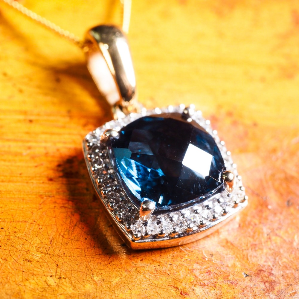 A cushion-cut deep blue topaz gemstone set in a square diamond halo pendant on a gold chain. View X