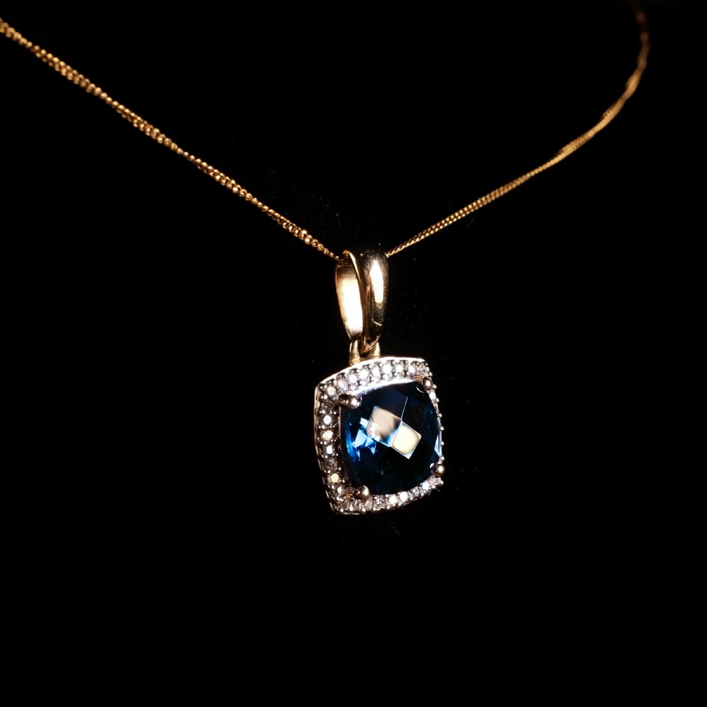 A cushion-cut deep blue topaz gemstone set in a square diamond halo pendant on a gold chain. View XI