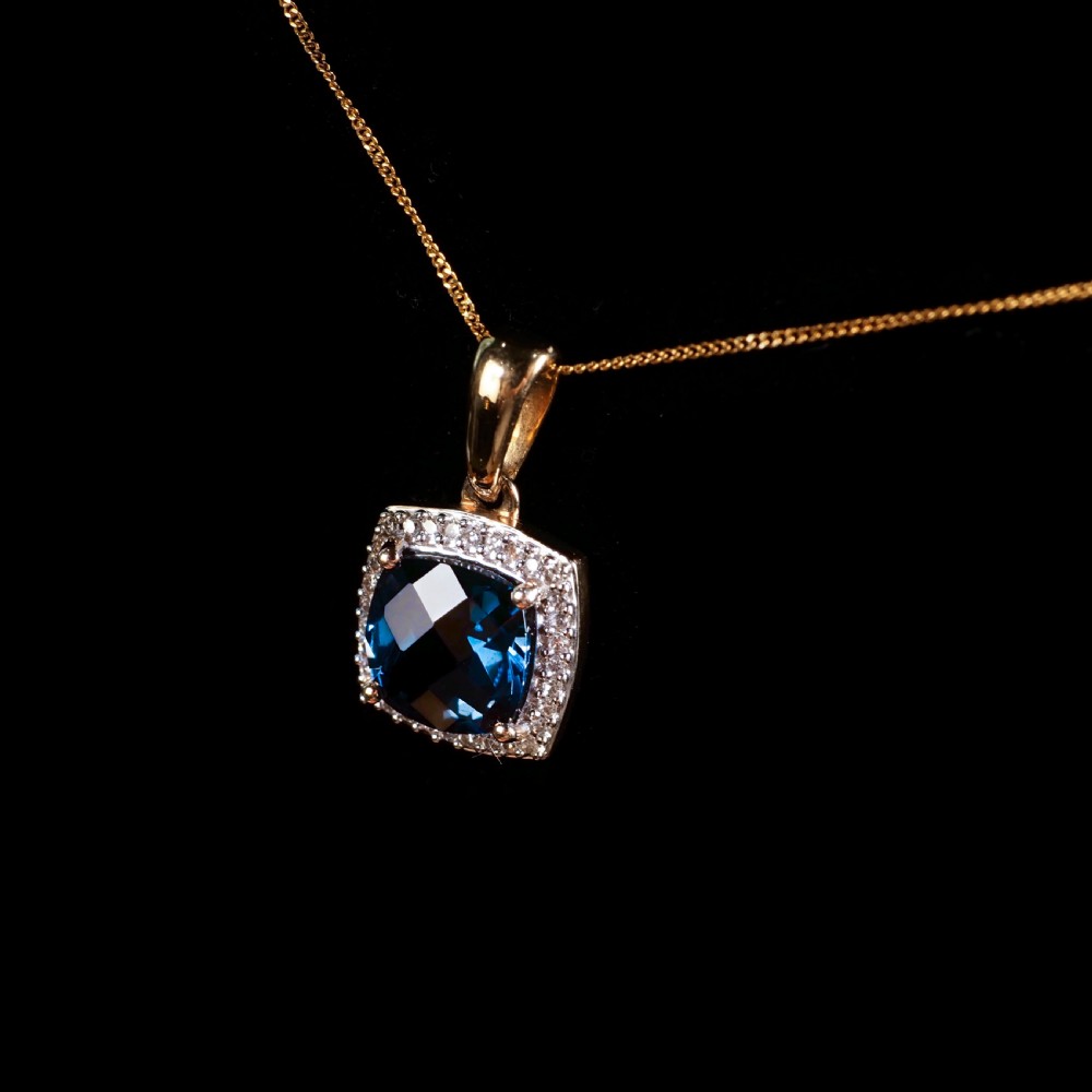 A cushion-cut deep blue topaz gemstone set in a square diamond halo pendant on a gold chain. View XII