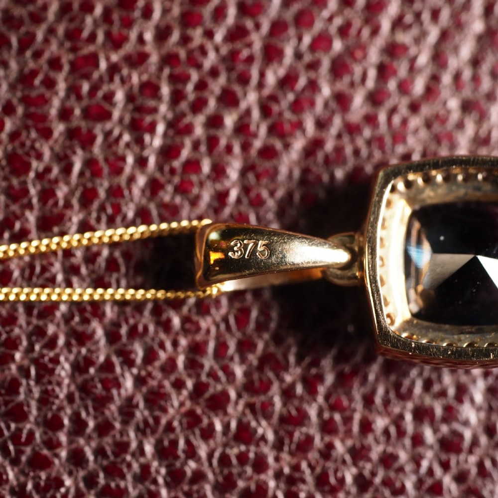 A cushion-cut deep blue topaz gemstone set in a square diamond halo pendant on a gold chain. View XIV