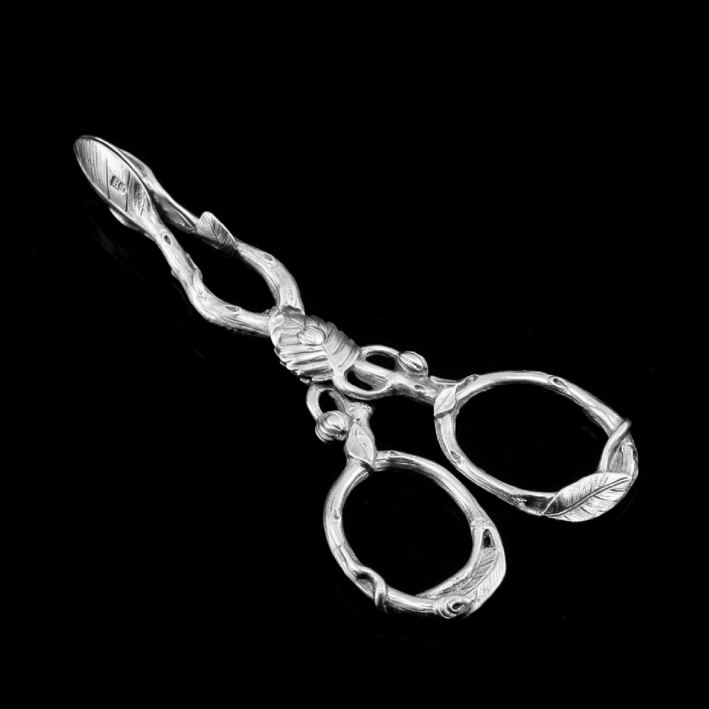 A pair of Victorian sterling silver scissor-action sugar nips crafted in a detailed branch and leaf design against a black background.
