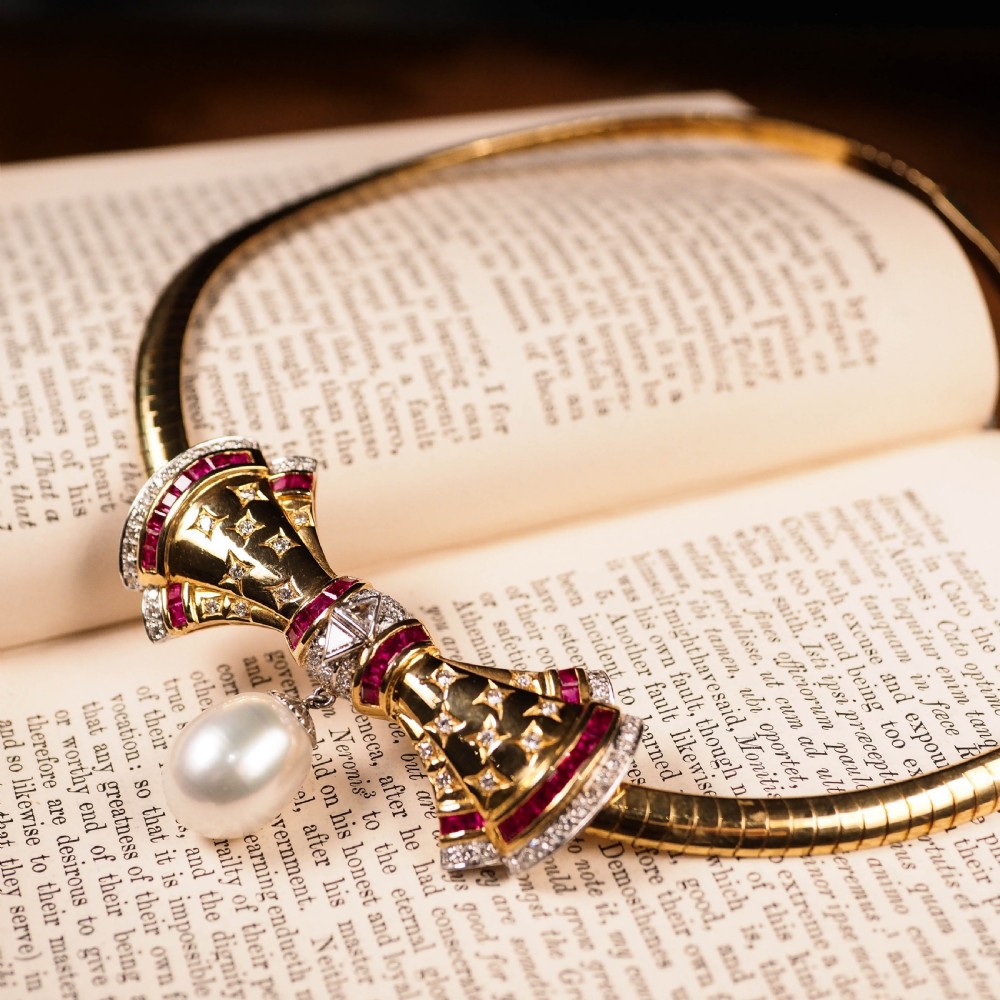 A structured 18k yellow gold collar necklace featuring a central bow motif accented with rubies and star-set diamonds above a teardrop pearl. View III