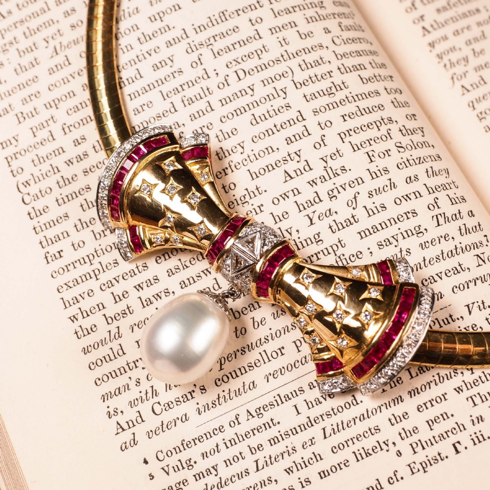 A structured 18k yellow gold collar necklace featuring a central bow motif accented with rubies and star-set diamonds above a teardrop pearl. View V