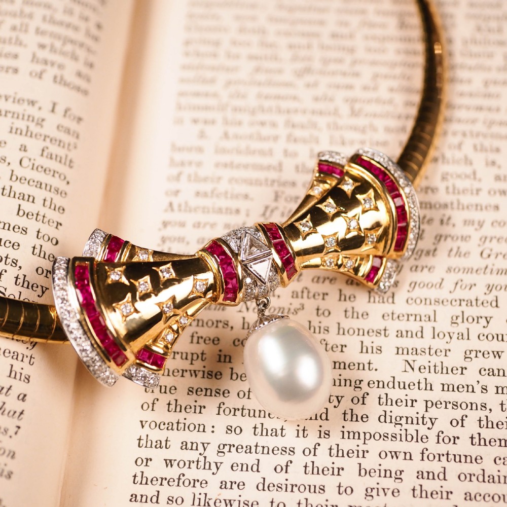 A structured 18k yellow gold collar necklace featuring a central bow motif accented with rubies and star-set diamonds above a teardrop pearl. View VI