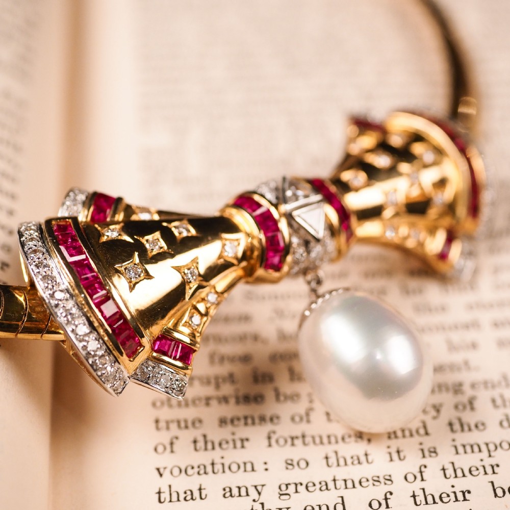 A structured 18k yellow gold collar necklace featuring a central bow motif accented with rubies and star-set diamonds above a teardrop pearl. View VII