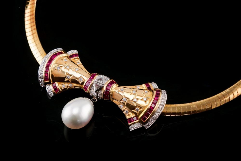 A structured 18k yellow gold collar necklace featuring a central bow motif accented with rubies and star-set diamonds above a teardrop pearl. View VIII