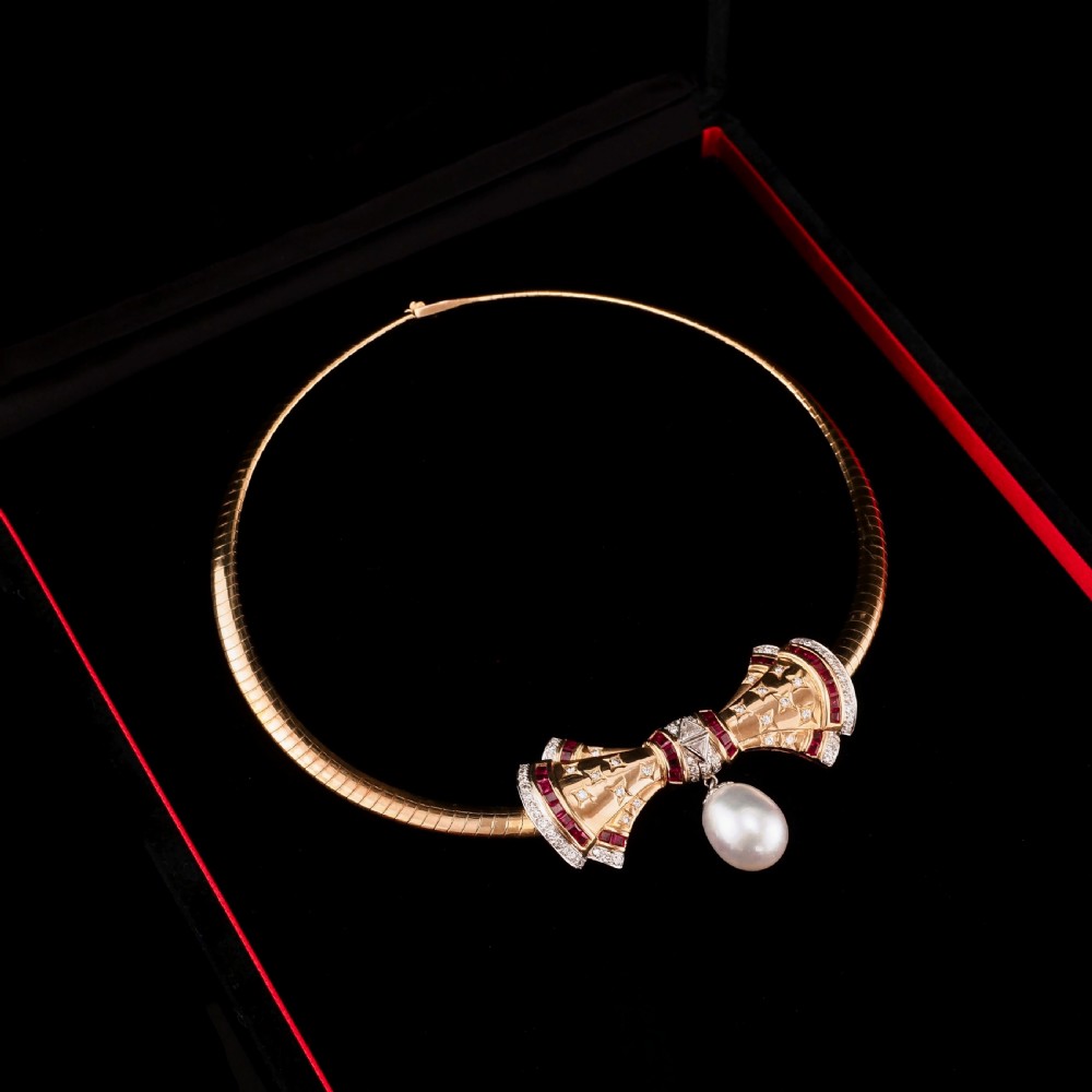 A structured 18k yellow gold collar necklace featuring a central bow motif accented with rubies and star-set diamonds above a teardrop pearl. View XXVI