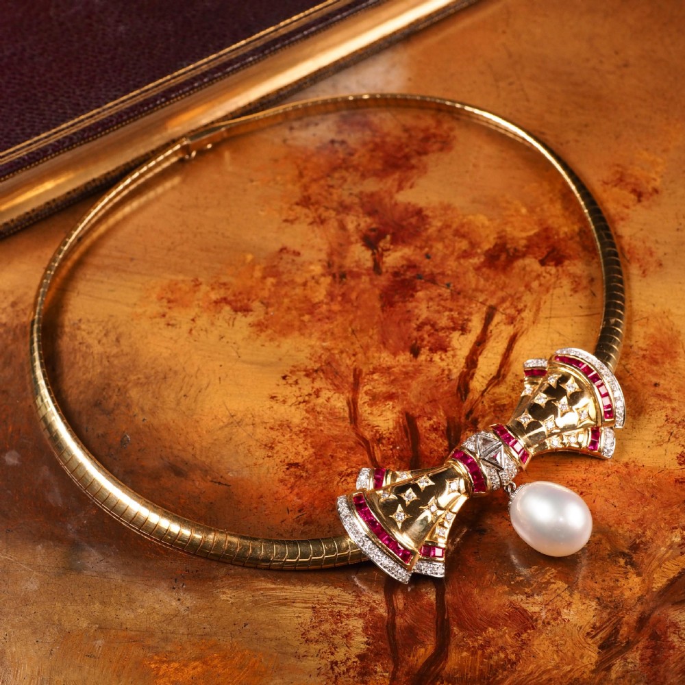 A structured 18k yellow gold collar necklace featuring a central bow motif accented with rubies and star-set diamonds above a teardrop pearl. View XXVII