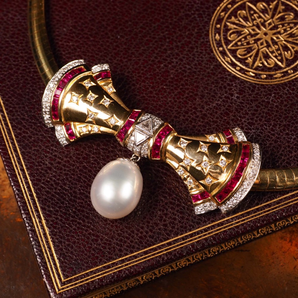 A structured 18k yellow gold collar necklace featuring a central bow motif accented with rubies and star-set diamonds above a teardrop pearl. View XXVIII