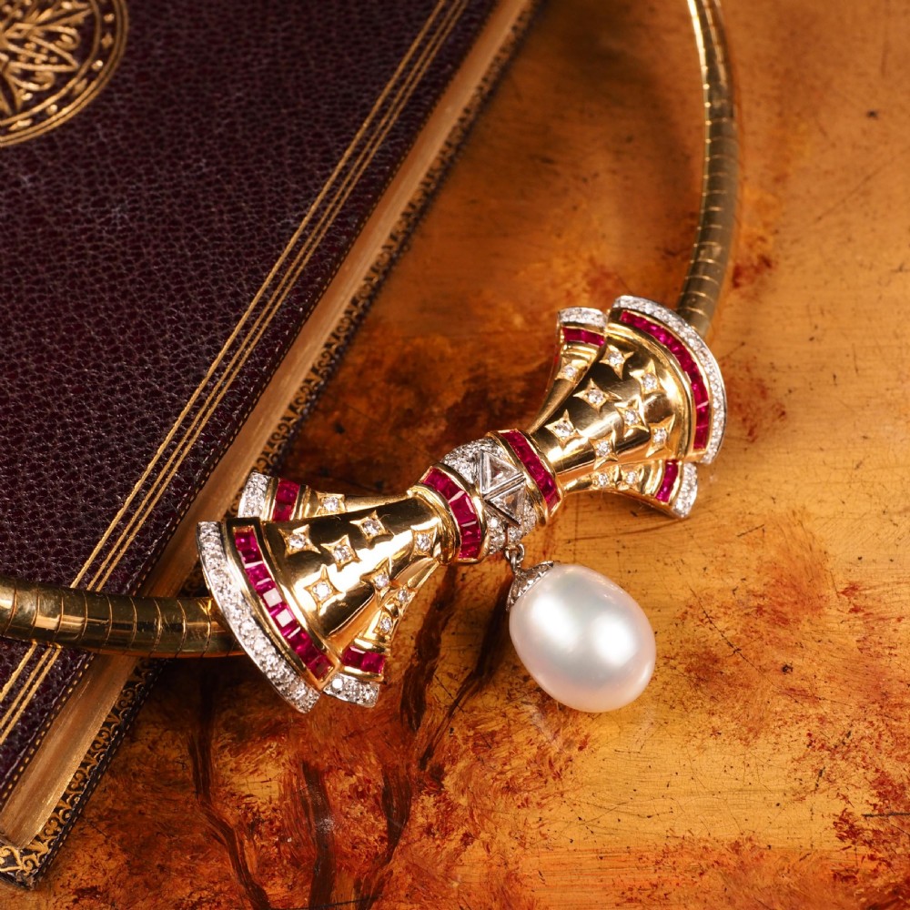 A structured 18k yellow gold collar necklace featuring a central bow motif accented with rubies and star-set diamonds above a teardrop pearl. View XXIX