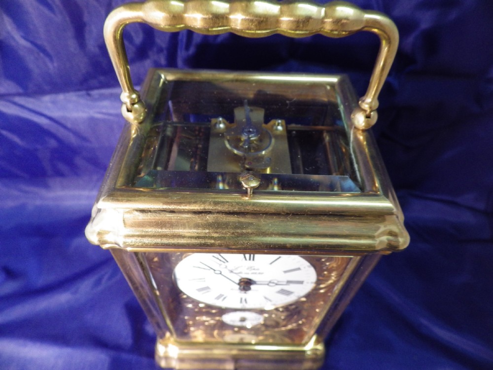 A rectangular brass repeating carriage clock featuring a top carrying handle, glass side panels, an engraved brass mask, and a white enamel dial with Roman numerals. View V