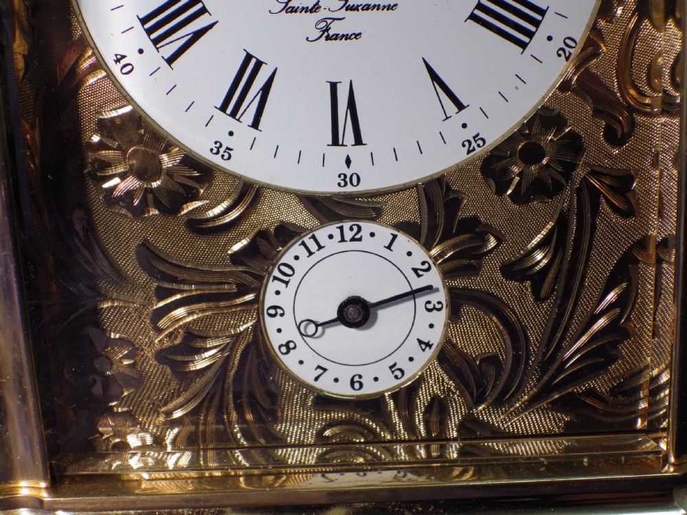A rectangular brass repeating carriage clock featuring a top carrying handle, glass side panels, an engraved brass mask, and a white enamel dial with Roman numerals. View VI