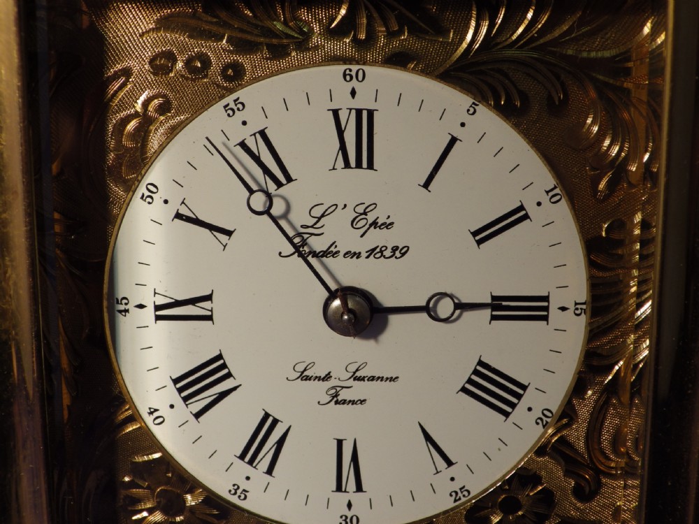 A rectangular brass repeating carriage clock featuring a top carrying handle, glass side panels, an engraved brass mask, and a white enamel dial with Roman numerals. View VII