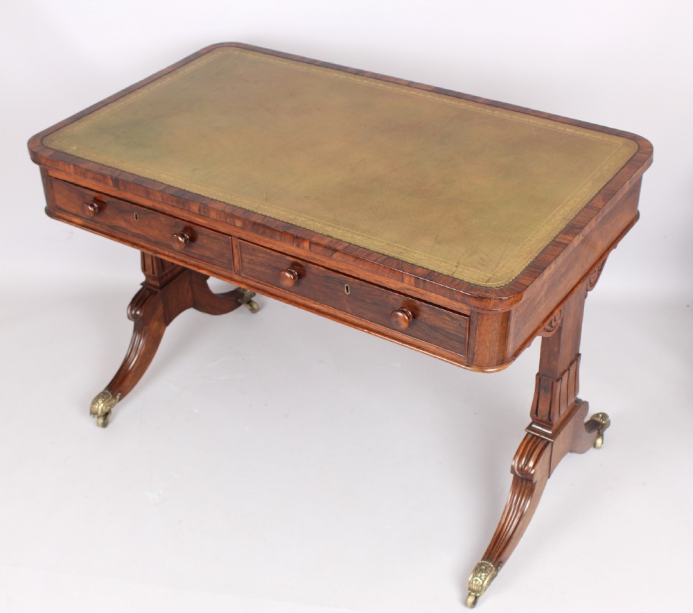 An antique rosewood writing table featuring a green leather inset surface, dual frieze drawers with wooden knobs, and splayed legs with brass paw castors. View IV