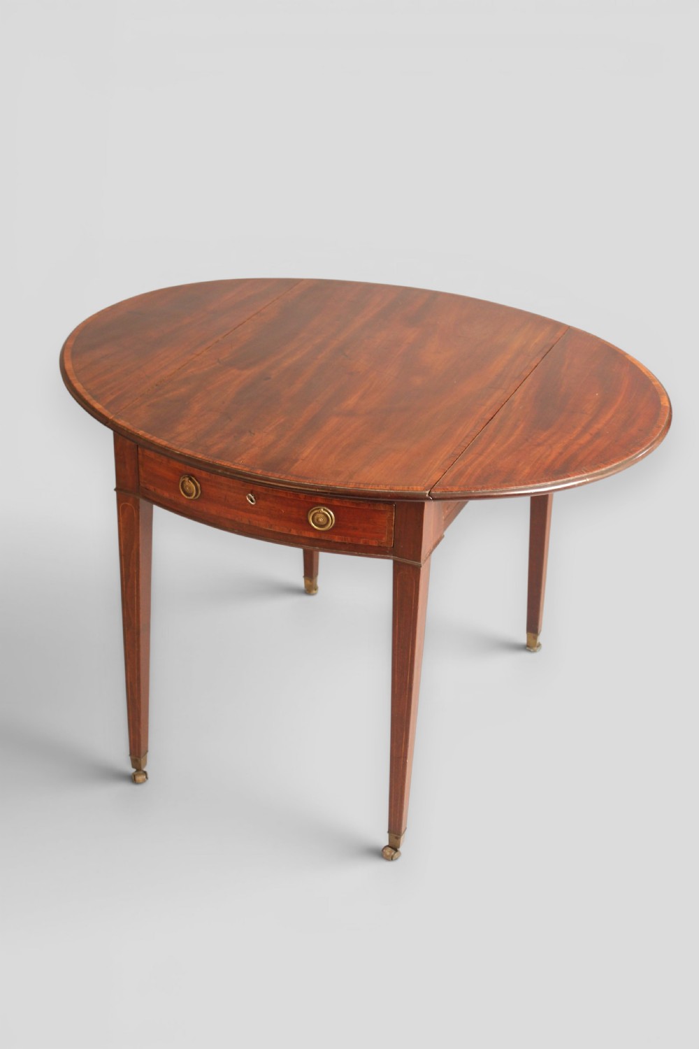 A George III period mahogany Pembroke table featuring an oval top with two drop leaves extended, a drawer with brass ring handles, square tapered legs with inlay, and brass castors.