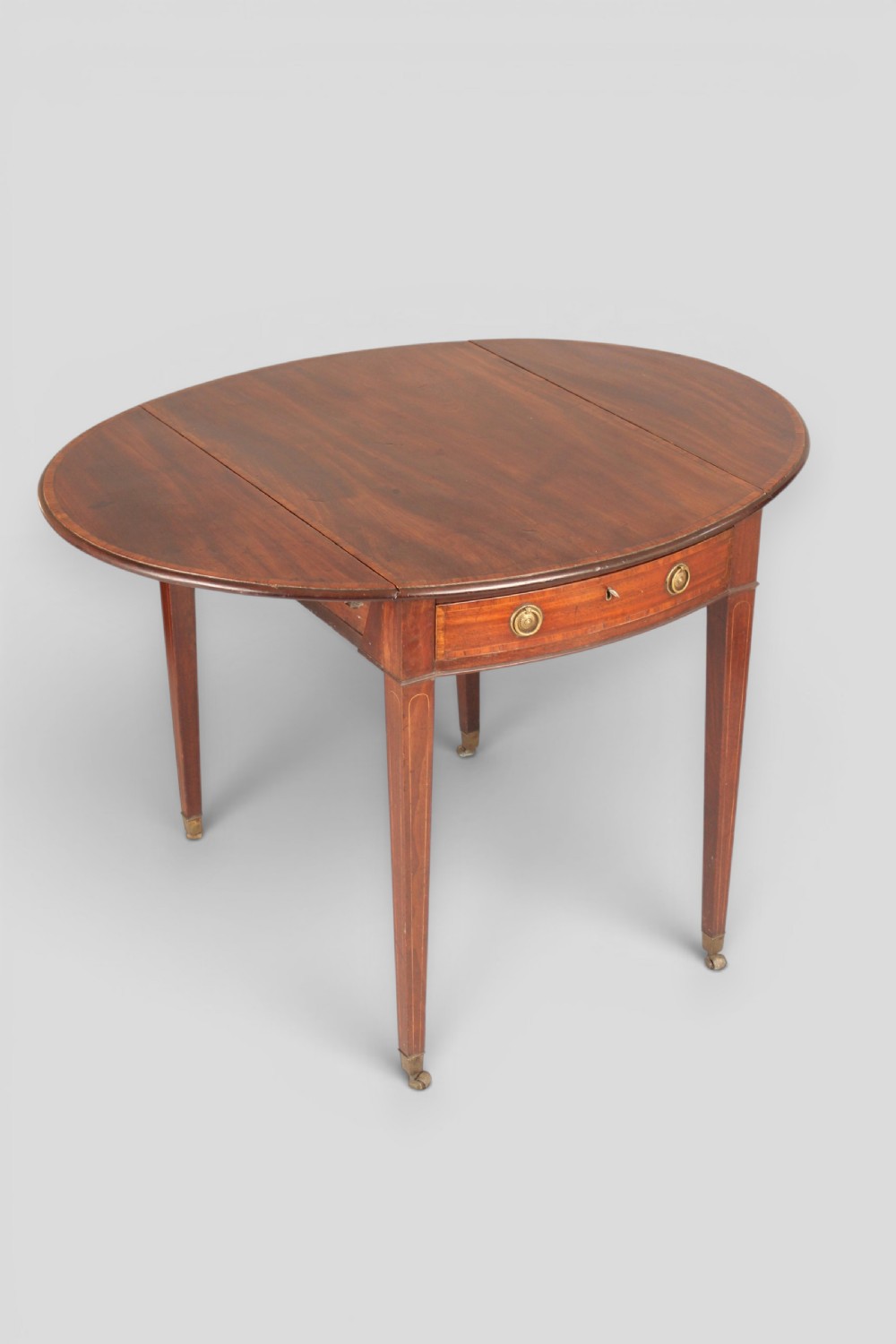 A George III period mahogany Pembroke table featuring an oval top with two drop leaves extended, a drawer with brass ring handles, square tapered legs with inlay, and brass castors. View II
