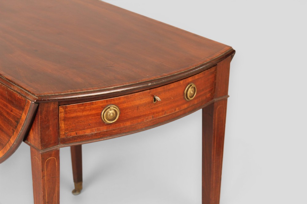 A George III period mahogany Pembroke table featuring an oval top with two drop leaves extended, a drawer with brass ring handles, square tapered legs with inlay, and brass castors. View III
