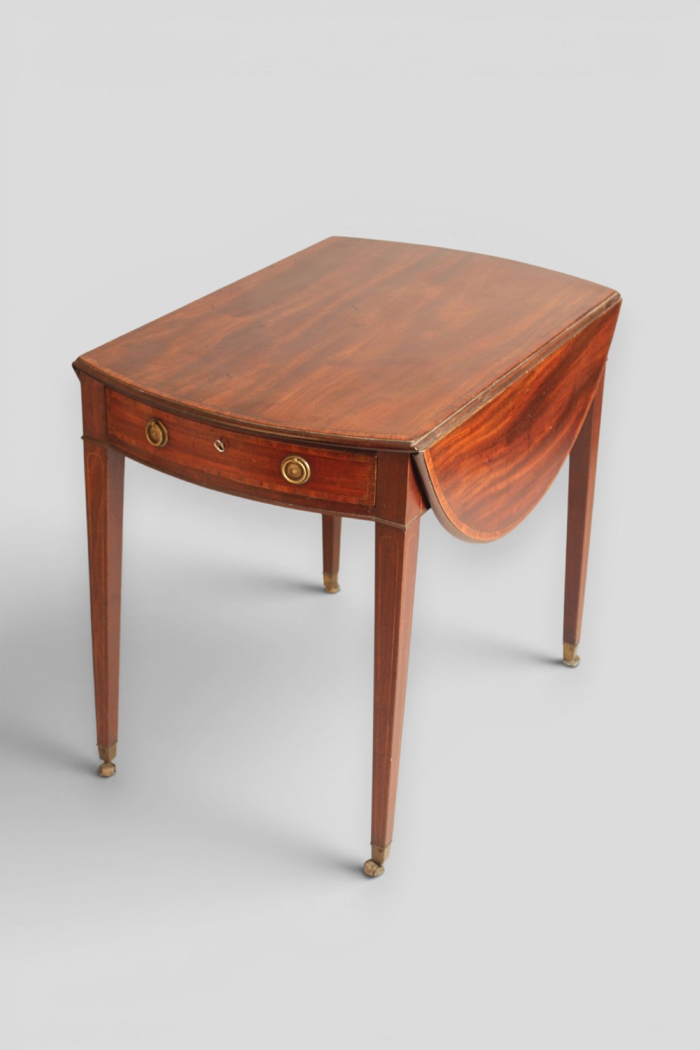 A George III period mahogany Pembroke table featuring an oval top with two drop leaves extended, a drawer with brass ring handles, square tapered legs with inlay, and brass castors. View IV