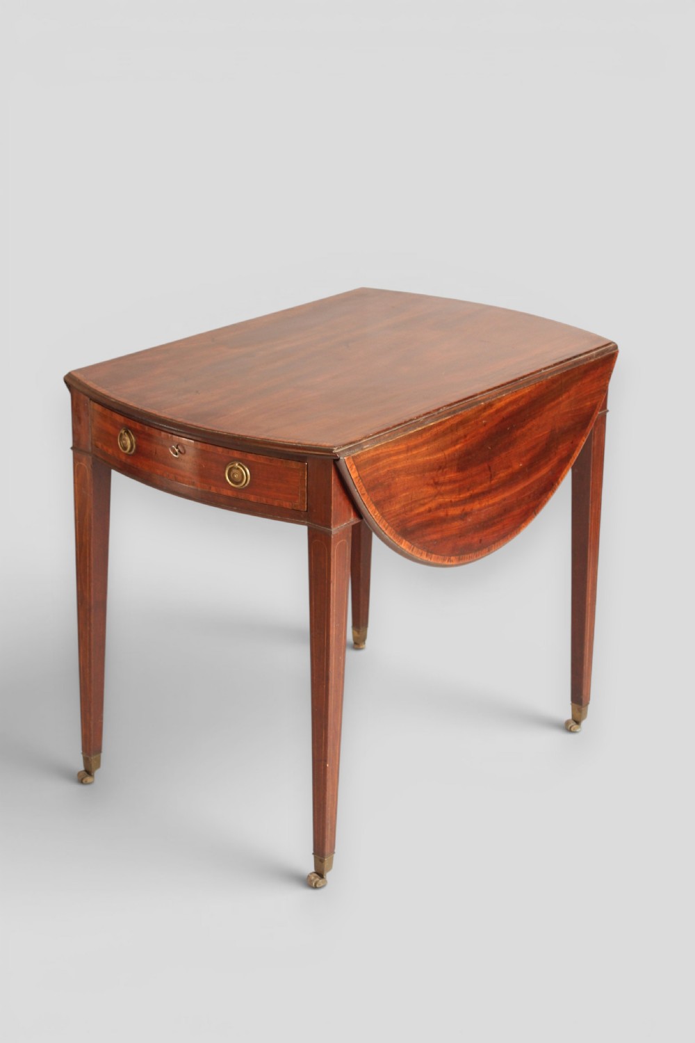 A George III period mahogany Pembroke table featuring an oval top with two drop leaves extended, a drawer with brass ring handles, square tapered legs with inlay, and brass castors. View V