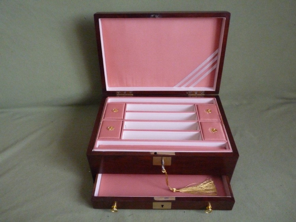 An open mahogany jewellery box featuring a pink-lined interior with tiered compartments, a lower drawer, and brass fittings.