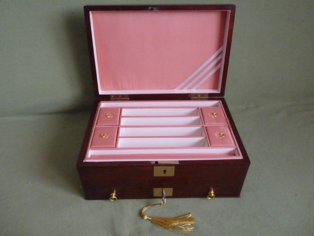 An open mahogany jewellery box featuring a pink-lined interior with tiered compartments, a lower drawer, and brass fittings. View III