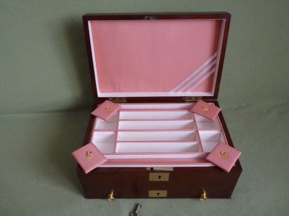 An open mahogany jewellery box featuring a pink-lined interior with tiered compartments, a lower drawer, and brass fittings. View V
