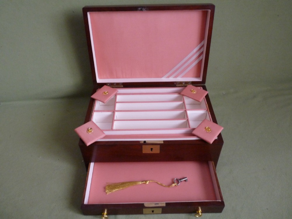 An open mahogany jewellery box featuring a pink-lined interior with tiered compartments, a lower drawer, and brass fittings. View VII