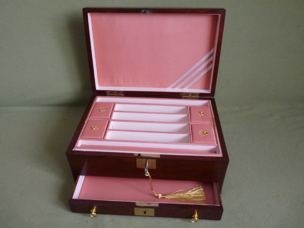 An open mahogany jewellery box featuring a pink-lined interior with tiered compartments, a lower drawer, and brass fittings. View X