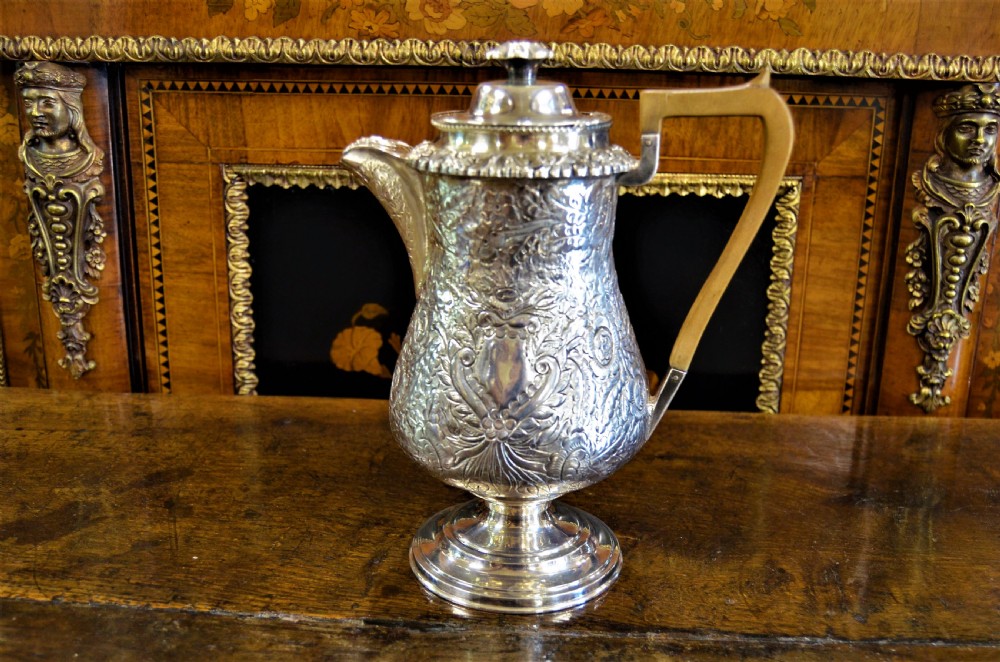 A Georgian silver-plated water jug featuring chased floral repousse scrollwork, a pedestal base, a hinged lid, and an angled timber handle.