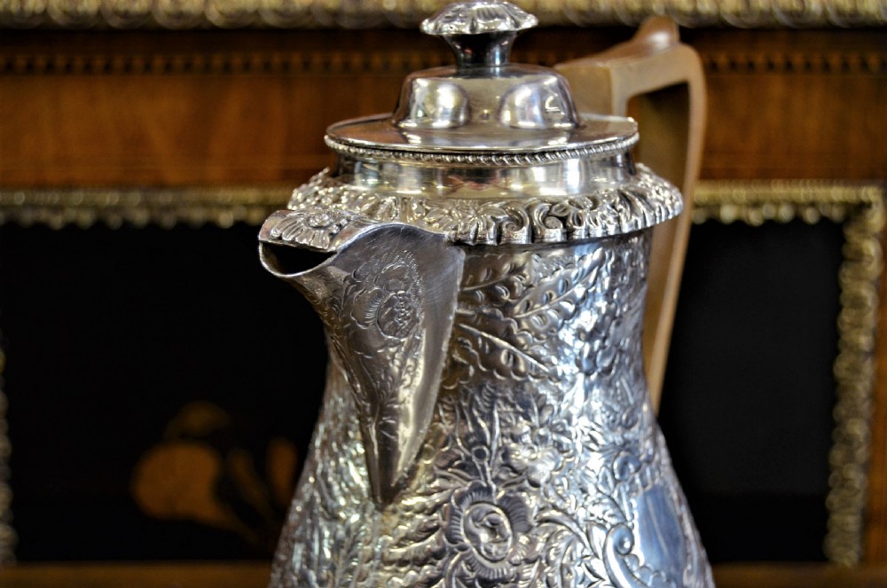 A Georgian silver-plated water jug featuring chased floral repousse scrollwork, a pedestal base, a hinged lid, and an angled timber handle. View II