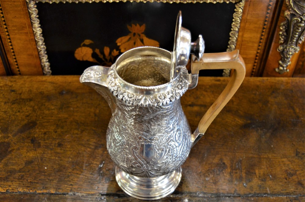 A Georgian silver-plated water jug featuring chased floral repousse scrollwork, a pedestal base, a hinged lid, and an angled timber handle. View V