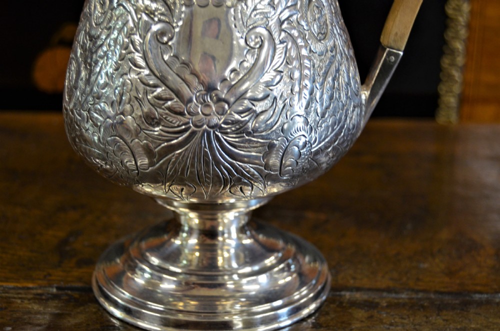 A Georgian silver-plated water jug featuring chased floral repousse scrollwork, a pedestal base, a hinged lid, and an angled timber handle. View VI