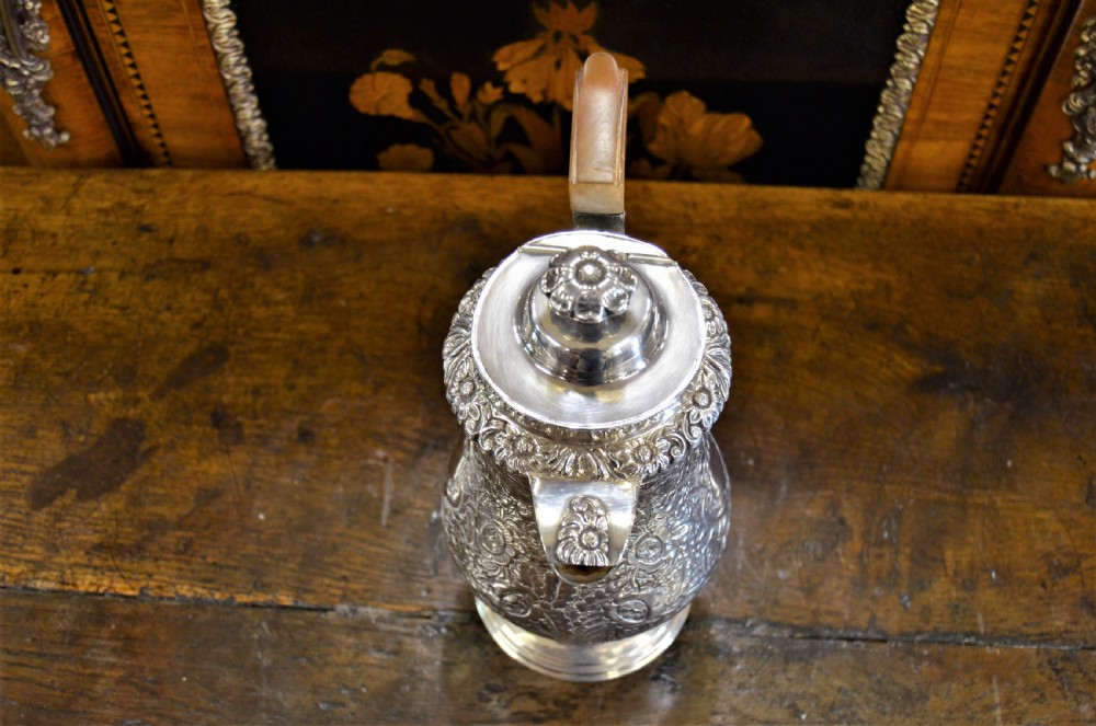 A Georgian silver-plated water jug featuring chased floral repousse scrollwork, a pedestal base, a hinged lid, and an angled timber handle. View VII