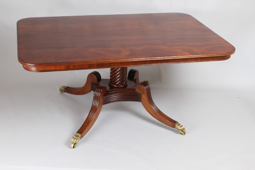 A George IV mahogany breakfast table featuring a polished rectangular top with rounded corners, supported by a spiral-carved pedestal and four reeded legs with brass lion paw castors.